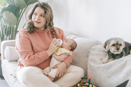 Young mother feeding a newborn baby with a bottle sitting on a couch with a small dog next to her